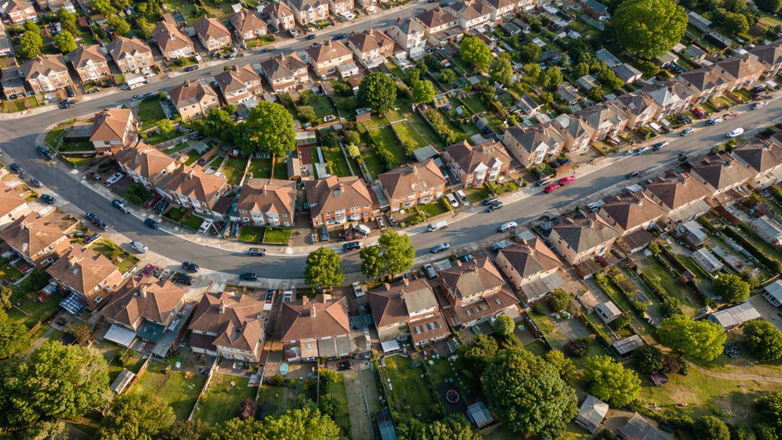 Aerial shot of a suburban neighbourhood showing rows of houses, green gardens, and tree-lined streets.